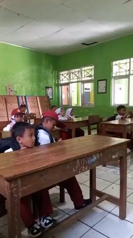The video shows a classroom with young students sitting at desks. The walls are painted green. In the foreground, two boys are visible at a wooden desk. The boy in the front is leaning over the desk with his mouth open, looking off to the right. The boy behind him is wearing a white shirt and a red hat, also looking to the right. Other students, also wearing red hats and uniforms, are visible in the background, seated at similar desks. Some students appear to be looking forward, possibly at a teacher or a whiteboard which is not in frame. The classroom appears to be sparsely decorated with some posters on the walls. The overall lighting suggests it is daytime.