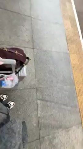 A woman with long auburn hair is standing on a train platform, filming a selfie video. She's wearing a gray speckled coat over a white top. Behind her, people with luggage are waiting to board the train, which is white with black accents. The platform has a gray tiled floor and a metal and glass roof. The video is filmed vertically and slightly shaky.