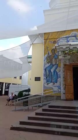 The video shows the exterior of a church. There are steps leading up to the entrance, which has large wooden doors. Above the entrance is a mosaic depicting religious figures. To the left of the entrance is a ramp. There are also white cloth canopies stretched over the area. In the background, there are buildings and trees. The sky is visible, and it appears to be daytime. The camera pans slightly to the right.