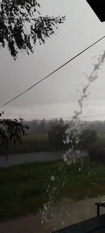 The video captures a view of a rural landscape during a rain shower. Water is seen pouring down from above, obscuring the view of a wire or cable that runs horizontally across the frame. The background reveals a body of water, possibly a river or pond, surrounded by green grass and trees. The sky is overcast, indicative of rainy weather. The overall scene depicts a serene yet potentially heavy downpour in an outdoor, natural setting.