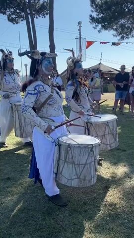 The video displays a group of three individuals dressed in white and blue costumes, playing large drums. They are positioned outdoors on a grassy area, with trees and tents in the background. The individuals are facing the viewer and appear to be performing a drumming ritual or parade. The costumes include elaborate headdresses adorned with feathers and what appear to be animal horns. Their faces are painted with blue and white markings. The drums are rustic in appearance, with wooden frames and weathered drumheads. Other people are visible in the background, some watching the performance and others walking around. The scene appears to be taking place during the daytime under a clear sky. The camera is positioned at a medium distance, capturing the drummers from the waist up, with some of the surrounding environment visible. The overall atmosphere suggests a festival or cultural event.