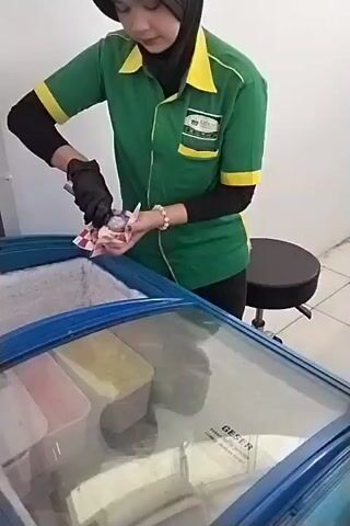A person wearing a green uniform with yellow accents, black sleeves, black gloves, and a black hijab is standing and scooping ice cream from a freezer. The freezer is blue with a glass lid. Inside the freezer are containers of ice cream and what appears to be packaging. The person is holding a scoop in their right hand and a checkered paper wrapper in their left hand, into which they are placing the scooped ice cream. The background is a plain white wall and a black stool.
