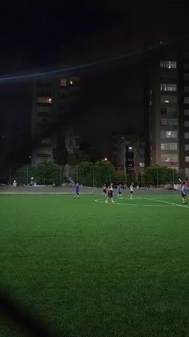 A group of people are playing soccer on a green turf field at night. The field is illuminated by lights, and there are tall apartment buildings in the background. One person in a white shirt and blue shorts is visible on the left side of the frame, walking across the field. Several other individuals are scattered across the field, some in blue and white uniforms. The sky is dark, indicating nighttime.