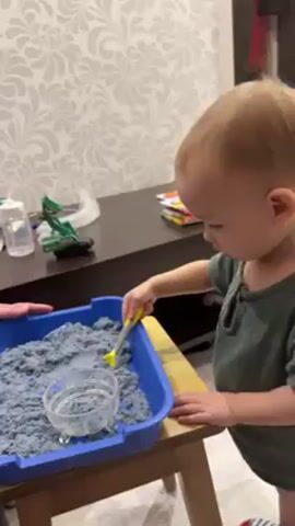 A young child is playing with blue kinetic sand in a blue plastic container. The container is on a small wooden table. The child, who is wearing a green shirt, is using a yellow and silver spoon to scoop the sand into a small, clear plastic bowl within the container. The child stands facing right, with their left hand resting on the edge of the table for balance. In the background, there is a dark brown dresser with a baby bottle, a green toy truck, and some books on top. The wall behind the dresser is covered with a white and grey patterned wallpaper. A hand is visible on the left side of the frame, possibly belonging to an adult who is supervising the child.