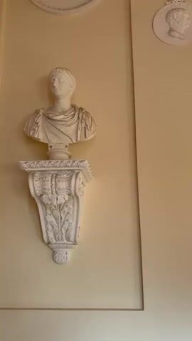The video pans across a room with pale yellow walls, decorated with various sculptures and artwork. On the left, a white bust of a Roman-style figure stands on an ornate white bracket mounted on the wall. To the right of this, several round white reliefs, each depicting a profile of a man with a crown, are mounted in a line. Further to the right, a full-length white marble statue of a nude woman stands on a pedestal, partially covered by drapery. The camera then moves to show a large framed painting of classical ruins hanging on the wall. Below the painting is an ornate wooden chest with gold detailing. Two chairs with red velvet upholstery and gold frames are positioned in front of the chest. The ceiling is visible at the top of the frame and features intricate molding and a chandelier. The floor is tiled in a dark and light pattern. The lighting appears to be natural, coming from an unseen source, casting soft shadows. The overall impression is of a well-decorated, possibly historical or museum-like interior.