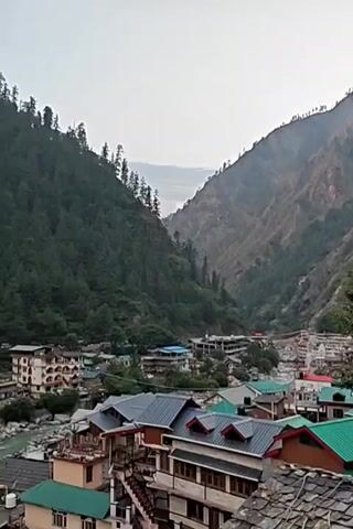 This image presents a panoramic view of a mountain town nestled in a deep valley, characteristic of a location like Manikaran, India. Towering mountains, densely covered with green coniferous trees, dominate the background and frame the scene on both sides. A light blue-green river flows through the bottom of the valley, winding its way past rocky banks.

Numerous buildings, appearing to be multi-story houses and perhaps hotels or guesthouses, are constructed along the riverbanks and climb up the lower slopes of the mountains. These structures exhibit varied architectural styles, some with flat roofs and others with distinctive sloping roofs, often in shades of green, grey, or brown. Many buildings feature balconies, offering views of the surrounding landscape.

The sky above is overcast, casting a soft, diffused light over the entire scene, suggesting either early morning, late afternoon, or a generally cloudy day. There are no people visible, and no specific activity is taking place. The overall impression is one of a serene and picturesque mountain settlement integrated with its natural, rugged surroundings. No discernible text is visible within the image.