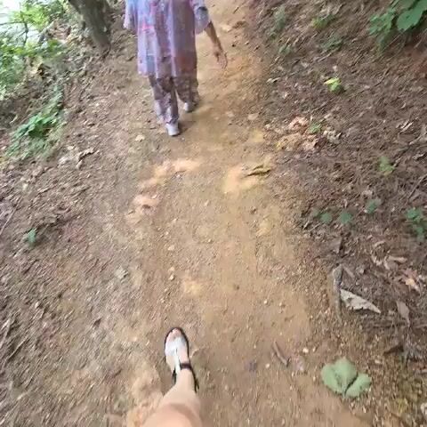 The video shows a person walking on a dirt path in a wooded area. The person is wearing a patterned robe and appears to be an elderly woman. Another person, who is further ahead on the path and mostly out of frame, is seen walking away from the camera. The path is lined with trees and dry leaves. The camera perspective is from behind the first person, implying the video is recorded by someone following them.