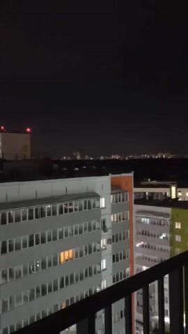 The video shows a night view from a balcony. In the foreground is a balcony railing and several apartment buildings. The buildings are multi-story and have lit windows. Beyond the apartment buildings, the city skyline is visible, with lights scattered across the horizon. The sky is dark with some visible stars. The video appears to be taken with a handheld device, as there is some slight movement.