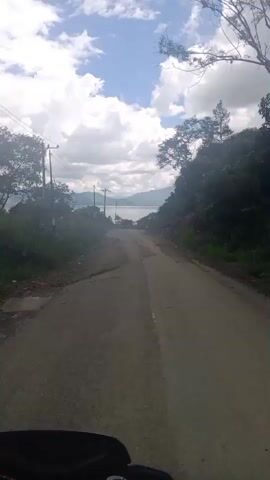 The video shows a first-person perspective of someone riding a motorcycle on a dirt road. The road is rough and uneven, with loose gravel and dirt. On either side of the road are steep, tree-covered hillsides. In the distance, a body of water, possibly a lake or the sea, can be seen. The sky is blue with scattered white clouds. There are power lines and utility poles visible along the side of the road. At approximately 00:04, another motorcycle can be seen riding in the distance on the same road. The rider's hands are visible at the bottom of the frame, gripping the handlebars. The video is shaky, indicating movement.