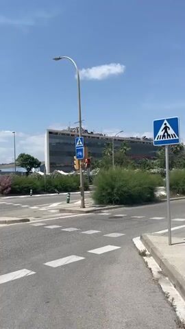 The video shows a first-person perspective of someone riding a scooter on a sunny day. The road is asphalt, with white crosswalk lines. On the left side of the road, there are bushes and a building in the distance. There is a traffic light with a pedestrian crossing sign. As the scooter moves forward, a small building with graffiti comes into view on the right, along with parked cars and a van. The sky is blue with scattered clouds.