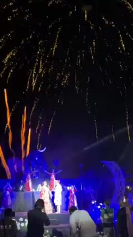 A wide, low-angle shot captures an outdoor wedding celebration at night, illuminated by stage lights and fireworks. A wedding party of about seven people is situated on a raised stage decorated with flower arrangements and crescent moon props. The couple stands center-stage, the bride wearing a red and gold traditional Indian dress, and the groom a white sherwani. Fireworks erupt behind them, creating a spectacle of vertical lines and a large, exploding starburst pattern. The ground level shows numerous guests in casual attire, many holding phones to record the fireworks. The stage is framed by white flower arrangements on either side of the walkway leading up to it. The atmosphere is celebratory and filled with visual spectacle.