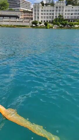The video is a point-of-view shot from a boat moving across a turquoise lake. The water has small ripples on the surface. On the left side of the frame, a large white building with many windows and balconies is visible, situated on a hillside covered with trees. Further to the right, the shoreline is lined with more trees and some smaller buildings. The sky is blue with scattered white clouds. The camera pans slowly to the right, revealing more of the tree-covered hillside and the lake. There are no people visible in the frame, nor are there any explicit interactions. The overall scene depicts a tranquil natural environment with a prominent man-made structure.
