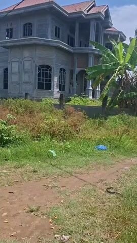 The video shows a two-story house under construction, appearing unfinished with open window spaces and a grey exterior. The house has a tiled roof and a veranda supported by white pillars. The house is surrounded by overgrown vegetation and a dirt path. There is a banana tree to the right of the house. A blue piece of trash is visible on the ground. The sky is clear and blue.