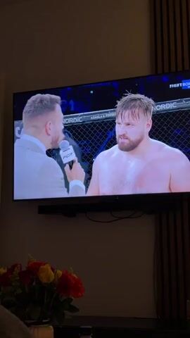 A large flat-screen TV displays an interview with a shirtless male fighter inside a cage, likely after a combat sports event. The fighter, with a beard and wet hair, is smiling broadly as he speaks into a microphone held by an interviewer in a light-colored suit jacket. The microphone has "FCR" written on it. In the background on the TV, parts of text like "A NORDIC," "KÄNSLA & FO," and "FIGHT BOX HO" are visible on the cage fencing. Below the TV is a soundbar.

In the foreground, a vibrant bouquet of red and yellow roses sits in a white vase on a table. Next to it, a clear plastic water bottle and a lit candle in a glass holder are visible. The scene appears to be an indoor living room setting, likely in the evening or at night, with a wooden slatted wall panel visible on the right. A soft, dark fabric, possibly part of a sofa or blanket, is in the bottom left corner.