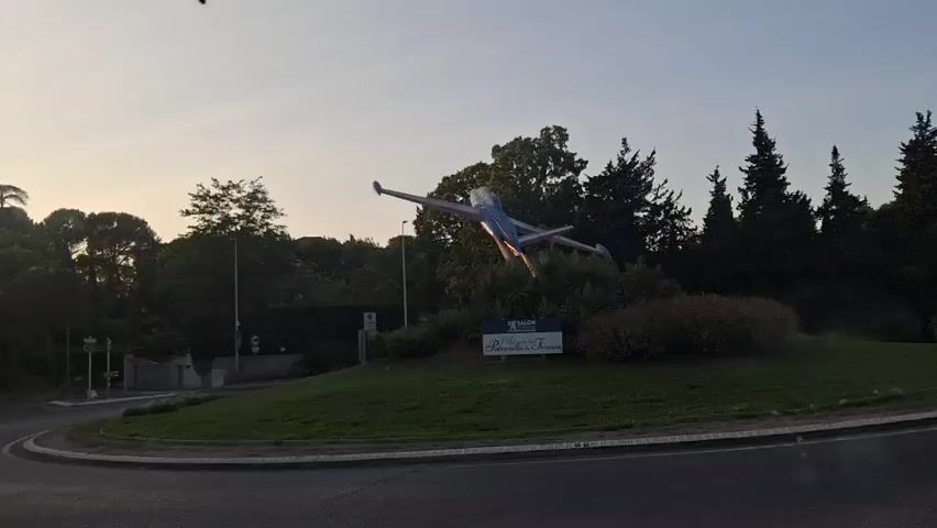 A statue of a small airplane is perched on a grassy roundabout. The plane is white and blue and appears to be in mid-flight, tilted upward with its wings extended. Behind the plane, there are trees and bushes. A sign with text in French is visible at the base of the statue. The sky above is a pale orange, suggesting either sunrise or sunset. The roundabout itself is paved and curves out of view.
