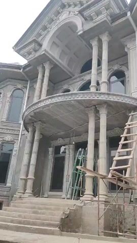 The video shows the exterior of a large, unfinished building with neoclassical architectural features. The facade is mostly gray with white decorative elements, and there are several windows with dark frames. A prominent balcony supported by columns is visible above the entrance, which is accessed by a set of stairs. Ladders and scaffolding are present around the building, indicating ongoing construction or renovation work. No people are clearly visible in the frame. The sky is overcast, suggesting a cloudy day.
