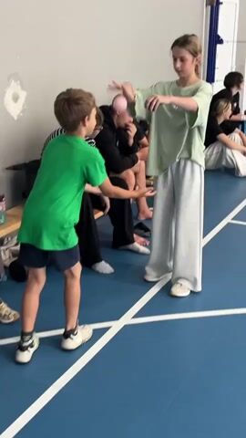 A group of young people are in a room with a blue floor marked with white lines, likely a gymnasium. In the foreground, a boy wearing a green t-shirt and navy shorts is facing a girl in a light green t-shirt and wide gray pants. The boy is in a throwing stance, reaching out with his left hand. The girl is also reaching out with her hands. A pink ball is in the girl's right hand, and she appears to be speaking. Behind them, several other people are sitting on a bench. One person wears a black and white striped shirt, another a black hoodie, and another person with a shaved head is visible. The environment appears to be an indoor recreational space.