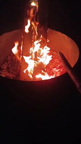 A close-up, top-down shot of a fire pit shows logs burning. Flames flicker and dance between two logs that are positioned vertically in the center of the pit. Embers and ash are visible at the bottom and sides of the pit. A stick is positioned diagonally from the bottom right to the center of the frame, touching one of the logs. Sparks fly upwards and to the right of the frame. The background is dark.