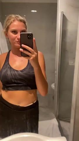 A woman stands in front of a mirror in a bathroom, taking a selfie. She is wearing a black sports bra and a black skirt. The bathroom has a sink, a toilet, and a shower. The walls are tiled with a marble pattern.