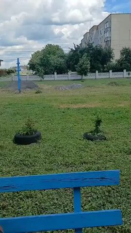 The video shows an outdoor scene. In the foreground, a blue wooden bench is visible. The camera pans across a grassy area with several small trees and bushes. Some of the plants are in tires that have been repurposed as planters. In the background, there is a white fence, a tall apartment building, and other houses. There is also a small, white statue of a person under a blue and gold canopy with crosses on top. The sky is partly cloudy.