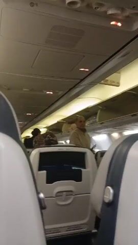 The interior of an airplane is shown. The airplane cabin is white with overhead compartments. Passengers are putting their luggage into the compartments. A man in a tan shirt is walking towards the back of the plane. The airplane seats are white and blue. The video is taken from the perspective of someone sitting in one of the airplane seats.