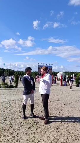 The video shows two men in an outdoor sandy arena under a bright blue sky with scattered clouds. The man on the left is dressed in equestrian attire, including a dark jacket, white breeches, black boots, and a black helmet. He wears a sash across his chest that appears to have some lettering or insignia on it. The man on the right is wearing a white shirt, dark pants, a brown hat, and a red scarf. He is holding a microphone and appears to be interviewing or speaking with the equestrian.

In the background, there are several people, some trees, and what looks like banners or flags. Further in the distance, there is a large screen or banner with images on it, and some tents or structures. The overall setting suggests an outdoor event, possibly a horse show or competition, given the equestrian attire and the arena.

The time of day appears to be daytime, likely morning or afternoon, judging by the bright sunlight and the shadows. The weather seems pleasant, with a clear blue sky. The atmosphere seems to be one of an organized event. There is no discernible text that can be clearly read in the video.