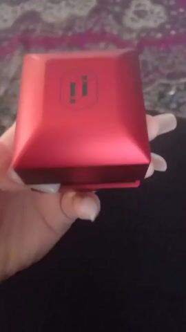 A hand with long, light-colored artificial nails opens a small, red, rectangular jewelry box. The box has a raised, faceted lid with a black logo featuring two stylized "i"s. Inside the box, resting on a black velvet lining, is a silver necklace with a delicate chain and a pendant shaped like a star or flower with multiple pointed petals, adorned with small, reflective stones. The lid of the box is fully open, revealing the necklace. The background is blurred, showing hints of purple and dark fabric.