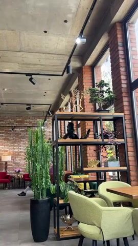 The media file shows an indoor scene with a contemporary design, likely a cafe or restaurant, in Adzhiryuara, Abkhazia. The space features exposed concrete ceilings, brick walls, and large windows that let in natural light, suggesting it's daytime. The decor is a mix of modern and natural elements.

Prominently displayed on a black metal shelving unit are several decorative objects. On the top shelf, two golden deer figurines with intricate, tree-like antlers stand next to a bonsai tree. The middle shelf holds abstract black sculptures, possibly made of wood or metal, and some greenery. The lower shelves display more plants, including a tall cactus in a black pot on the left, and a golden coral-like sculpture.

In the background, glimpses of seating areas are visible. A person can be seen sitting at a table on the left, partially obscured by a large potted plant. Two red chairs are also visible in the distance, adding a touch of color to the scene. The overall atmosphere appears relaxed and stylish, with a focus on bringing natural elements into the interior design. The lighting seems to be a combination of natural light from the windows and modern track lighting fixtures on the ceiling. No text is visible in the media file.