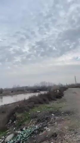 The video shows a view of a river from a muddy embankment.  There are trees in the background, and the sky is overcast but brightening.  The camera pans slightly along the river, then a young man wearing a cap and vest steps into frame for a brief moment, smiling at the camera.  The video ends with a view of the river and sky.
