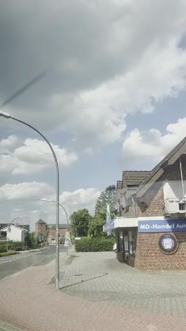 The video captures a street scene from a moving vehicle. On the right side of the frame, a brick building with a blue and white sign is visible, possibly a store. Further down the street, a white car is parked. The street has a sidewalk with red paving stones, and a bike lane is marked in red on the road. Streetlights are present along the sidewalk. Trees and residential houses with tiled roofs are seen on the left side of the street, interspersed with greenery. The sky is partly cloudy with a mix of white cumulus clouds and blue sky. The camera appears to be mounted at a slight angle, offering a perspective of the passing environment. The video is short and seems to be a continuous shot without any apparent edits or changes in camera angle.