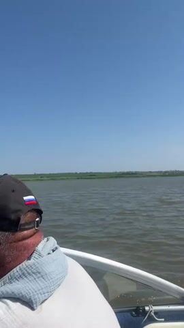 A video shows a person in a boat on a lake. The person is wearing a blue life jacket. The boat is moving, creating a wake. The sky is blue and clear. The shoreline is visible in the distance.