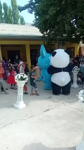 A group of people are gathered outdoors in front of a building. Several children are dancing and interacting with two costumed characters, a blue bunny and a panda. There are white pedestals with floral arrangements placed around the area. In the background, a car is parked and more people are observing the festivities. To the right, there is a structure draped with red fabric, suggesting a decorated area for an event. The environment appears to be a courtyard or open space adjacent to a building with a yellow facade and a tiled roof, surrounded by trees.