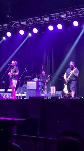 A live music performance on a stage with purple lighting. Three male musicians are visible. The musician on the left, wearing shorts and a t-shirt, sings into a microphone and plays a guitar. In the center, a drummer is seated at a drum set. To the right, a musician wearing a hat and shorts plays a guitar. They are performing on a stage with lighting rigs above and a dark backdrop behind them. The lower portion of the video shows a dark area, possibly an audience area, with silhouettes of people.