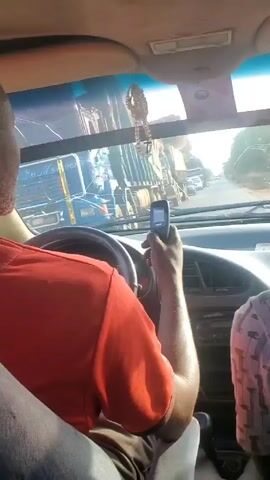 The video shows the inside of a car with a man in the driver's seat. He is wearing a red shirt and is on the phone while driving. The phone is a small, older model with buttons. The road ahead has other vehicles, including large trucks. There's a beaded ornament hanging from the rearview mirror. The dashboard of the car is visible, and another person is seated in the passenger seat, partially visible. The windshield has a crack in it.