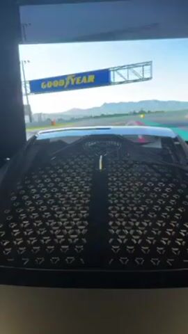 The video presents a view from inside what appears to be a driving simulator.  The car's hood is visible in the foreground, featuring a dark, triangular pattern on its surface. Through the windshield, a racetrack is displayed on a large screen, simulating the driving environment. A yellow marker is visible on the track. The sky is blue.  A mountain range can be seen in the distance.