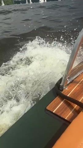 The video is taken from the perspective of a person on a moving boat. In the foreground, the wooden deck and a small portion of a tan seat are visible, along with a black rope. A clear windshield with a silver frame is also present. The boat is creating a wake in the dark, choppy water. In the background, numerous sailboats with white sails are dotted across a wide body of water. The horizon is lined with green trees, and the sky appears overcast with dramatic clouds. The overall scene depicts a day of sailing or a boat race on a lake or large body of water.