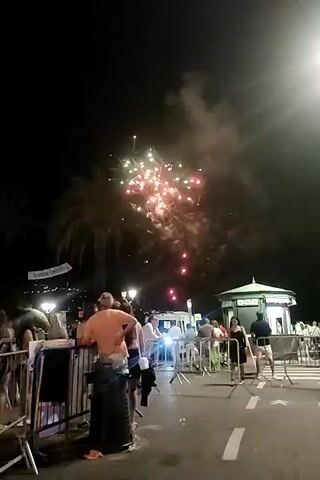 The video is a nighttime scene with fireworks in the sky. A crowd of people is gathered on a street, behind temporary barriers, watching the fireworks. The people are diverse in age and dress. Some are standing, some are leaning on the barriers. In the background, there are streetlights, buildings, and palm trees. A small green and white kiosk is visible on the right side of the frame. The fireworks are exploding in various colors, including red, green, and white, and are creating smoke plumes in the dark sky. The overall atmosphere appears festive.