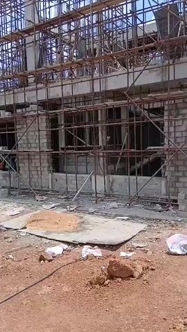 The video shows an exterior view of a multi-story building under construction. Scaffolding covers the entire structure, indicating extensive work is in progress. The building appears to be constructed with concrete and block walls, with many window openings visible. Debris, including sand, wood, and scattered white bags, litters the ground in front of the building, suggesting an active construction site. Several individuals, likely construction workers, are visible on different levels of the scaffolding and near the base of the building. The sky is overcast with some blue patches, and the overall environment suggests an outdoor setting during the daytime.