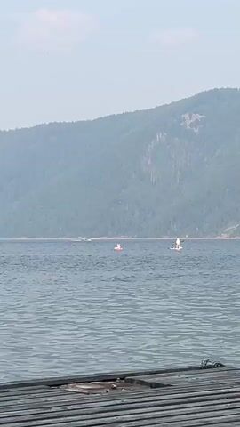A wide shot shows a body of water with a wooden dock in the foreground. In the middle ground, two people are on the water. The person on the left is on a small red inflatable raft. The person on the right is standing on a paddleboard. In the background, a heavily forested mountain rises steeply from the water. The sky is a hazy, light blue. The overall impression is a calm, outdoor scene.