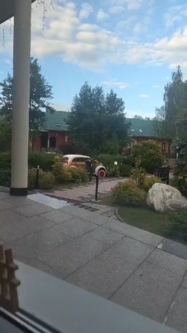 The video shows a view from a covered patio or porch area looking out onto a well-maintained garden and a building. A large, light-colored pillar is prominent in the foreground on the left. The ground in front of the porch is paved with large, grey square tiles.

Beyond the patio, the garden features lush green bushes and trees. A vintage, light orange car with a white roof and red wheels is parked on a paved area within the garden. A red and white object, possibly a fire alarm or a decorative item, stands next to the car. A large, light-colored boulder is visible in the right side of the garden, and a wooden bench is situated behind it.

In the background, a multi-story building with a green roof and red brick walls is visible. The building has several windows and architectural details. Trees surround the building and the garden.

The sky is blue with scattered white clouds, suggesting it is daytime, likely morning or afternoon. The overall impression is of a serene and pleasant outdoor setting, possibly a hotel, resort, or private residence in Цибино, Russia. No people are visible in the footage. There is no discernible text in the video. The scene appears calm and inviting.