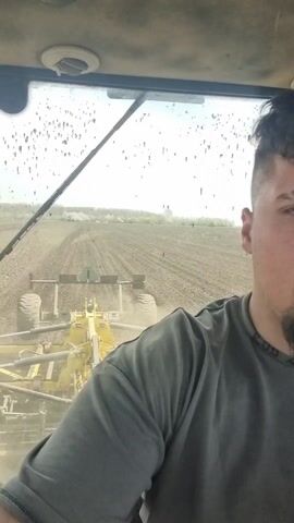 A man is operating a tractor in a field. The tractor is pulling a yellow piece of farm equipment that is tilling the soil. The man is wearing a gray shirt and has a modern haircut. The view is from inside the tractor's cab, looking out at the field. The window is somewhat dirty.