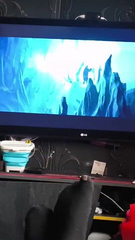 A pair of feet clad in dark socks rests in the foreground, with a small tear visible on the right big toe's sock, exposing the toenail. A large, black LG television is centered in the midground, displaying an animated scene predominantly featuring bright blue and white hues, depicting icy structures and dynamic abstract forms that rapidly shift. Below the television, on a dark surface, a light blue and white collapsible bucket, a black soundbar or speaker, and a small white desk calendar are visible among other indistinct items and cables. The background wall is dark with a subtle textured pattern.