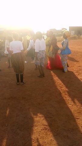 A group of people is standing and dancing in a dirt field, illuminated by a bright sun. Most of the individuals are wearing white tops with patterned skirts. There is one person in a red, feathered dress, and another in a blue and yellow outfit. The camera angle is from above.