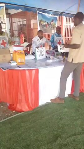 Here is a concise description of the video:

The video shows a scene taking place outdoors under a blue and white striped tent. Three men are the main subjects. Two of them are behind a table draped with a red and white tablecloth, wearing vests with the Red Cross emblem. One man in a white coat has a stethoscope around his neck. They appear to be engaging with the third man, who is standing on the other side of the table, reviewing documents in his hands.

Several objects are on the table, including posters with graphics of a tree and the Red Cross symbol, a laptop, and a yellow container with food aid written on it. A cardboard box with “ICRC” is also visible.

The ground beneath the tent is covered with artificial grass. In the background, there's a barred structure resembling a gate or window. There is also a banner hung with images of people.
