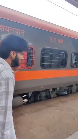 A man in a grey checkered shirt walks away from a train. The train is dark grey with orange stripes. The letters "NER" are visible on the side of the train. A second man in a blue t-shirt and black pants stands in the doorway of the train, holding the door open. The train is stopped at a platform. The platform is made of concrete tiles. There is a roof over the platform. The second man smiles and waves at the camera. The train has windows with bars. The number 264437 is visible on the side of the train. The word "SLEEPER" is also visible.
