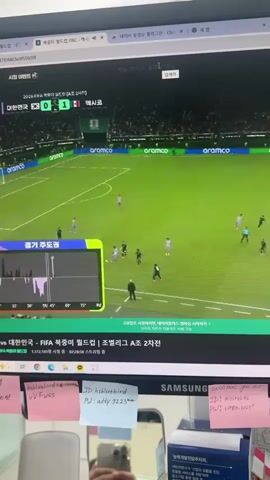 A live stream of a soccer match is displayed on a computer monitor. The screen shows a green soccer field with players in dark and light uniforms. A scoreboard indicates the score is 0-1. To the left of the field, a graph labeled "경기 모모모" is visible. The bottom of the monitor shows a desk with sticky notes attached. A small fan is positioned on the right side of the desk. The camera is focused on the monitor and the desk area.