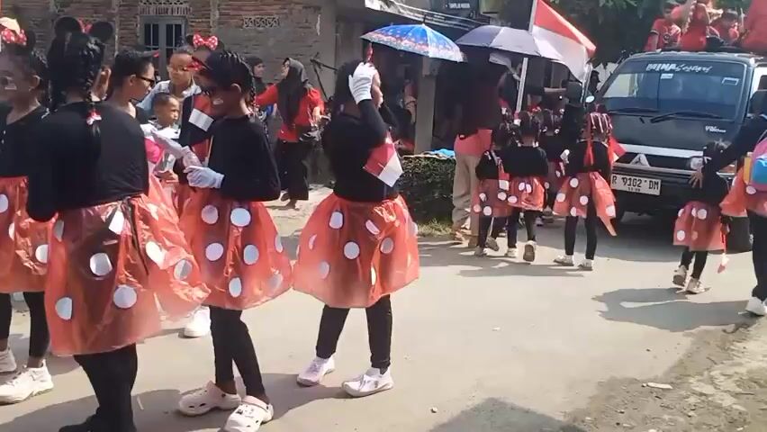 This is a video of a group of young girls in matching costumes walking in what appears to be a parade or procession. The girls are dressed in black long-sleeved shirts and red skirts with white polka dots, resembling Minnie Mouse costumes. They are also wearing white gloves and have red bows in their hair. Some of the girls are holding small umbrellas, and one girl is holding a miniature Indonesian flag. In the background, there are buildings, other people watching the procession, and a black van. The ground is paved, suggesting an outdoor setting. The overall atmosphere seems festive.