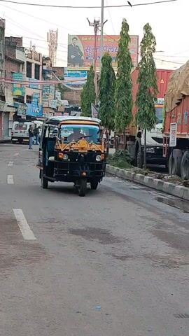A three-wheeled auto-rickshaw drives down a paved road. The auto-rickshaw is black and yellow, with a black tarp covering the passenger area. The driver is visible wearing a blue shirt. The auto-rickshaw has a large, light brown boulder strapped to its roof. In the background, there are buildings, utility poles, trees, and other vehicles, including a large truck. The road appears to be in an urban or semi-urban environment.