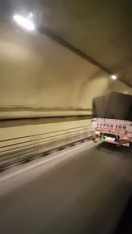 The video shows a truck driving through a tunnel. The tunnel has a beige-colored ceiling and walls, with lights spaced along the ceiling. There are guardrails on the left side of the tunnel. The truck is white with a gray tarp covering its cargo. The truck's taillights are visible and appear to be red, green, and blue. The video is shaky, suggesting it was filmed from a moving vehicle.