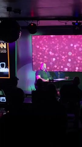A man is seated at a keyboard with a microphone in front of him, in what appears to be a dimly lit performance venue. He is looking upwards and speaking into the microphone. Behind him, a large screen displays a pink, bokeh-like pattern. To his left, a portion of a sign with yellow and black text is visible, reading "SUN" and "ON". Two disco balls hang from the ceiling, and green and purple lights illuminate the stage area. Silhouetted figures of an audience are visible in the foreground, facing the performer.